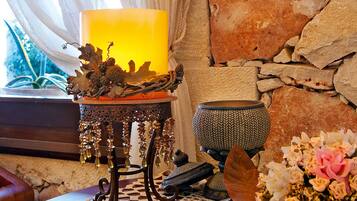 Uma vela sobre um suporte de madeira, com um parede de pedra ao fundo, uma janela com cortinas e um vaso com flores.