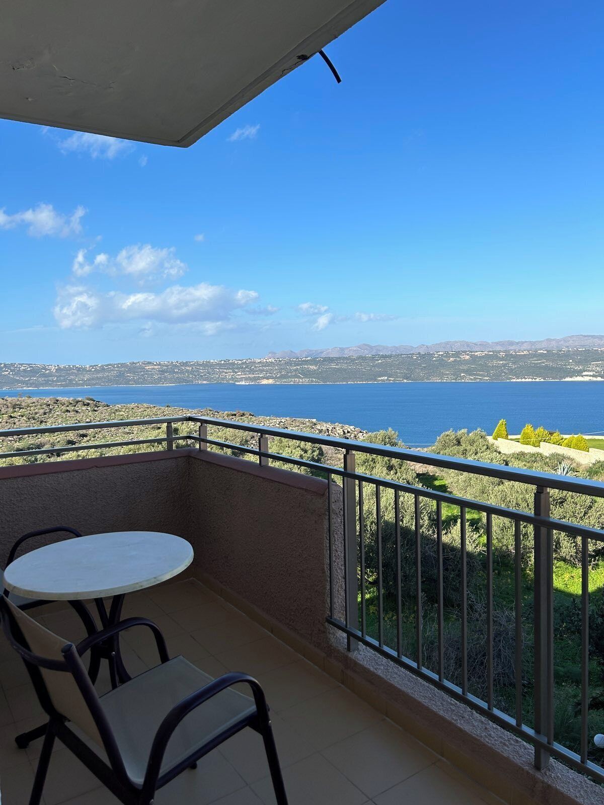 Balcón con una mesa y sillas, con vistas a un paisaje costero y una vista despejada del mar y colinas distantes.