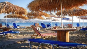 A beach with blue lounge chairs and straw umbrellas. - Areti Hotel (Chania)