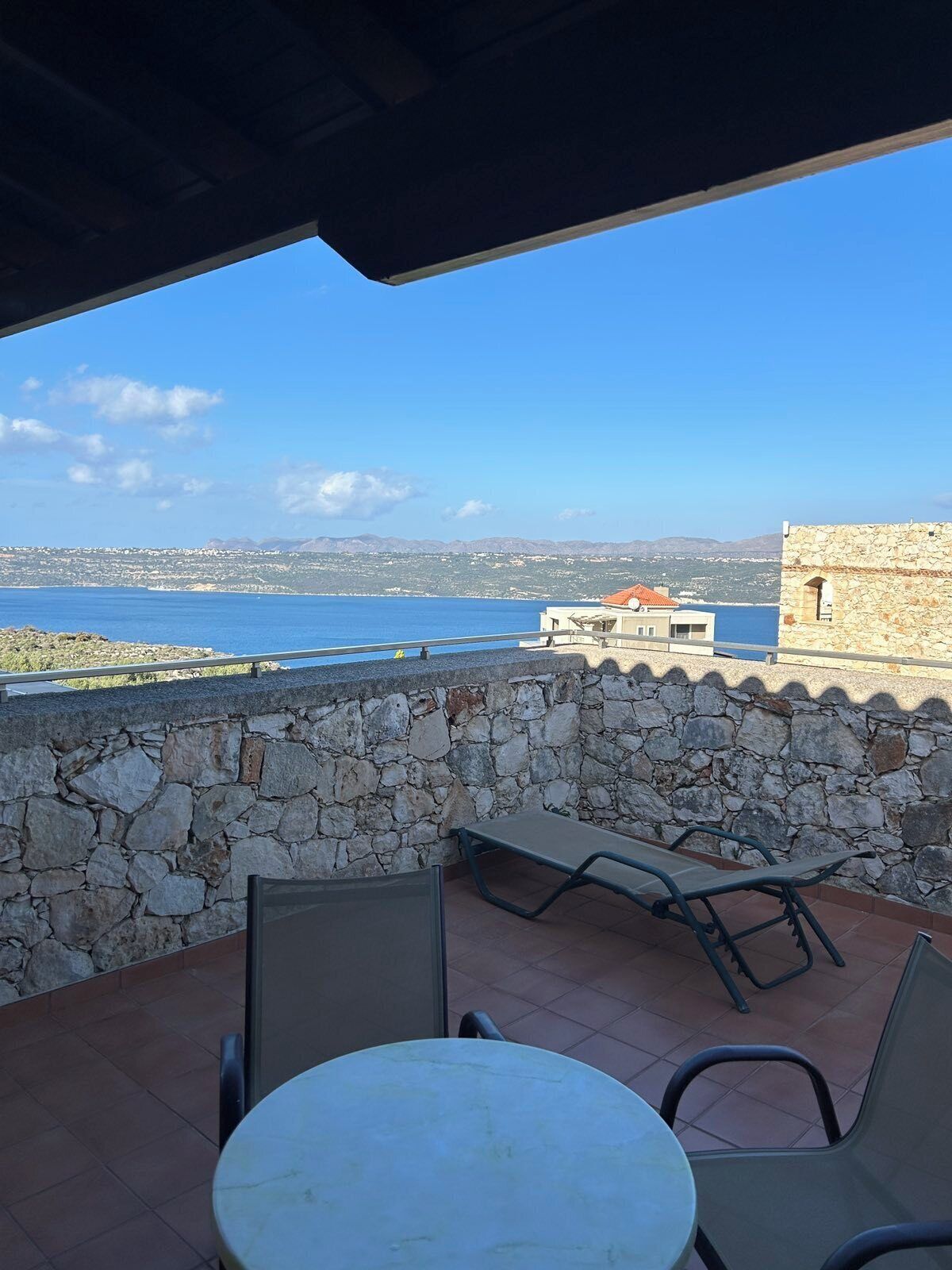 A terrace with a stone wall, a round table, and two chairs, overlooking a coastal view with a clear sky.