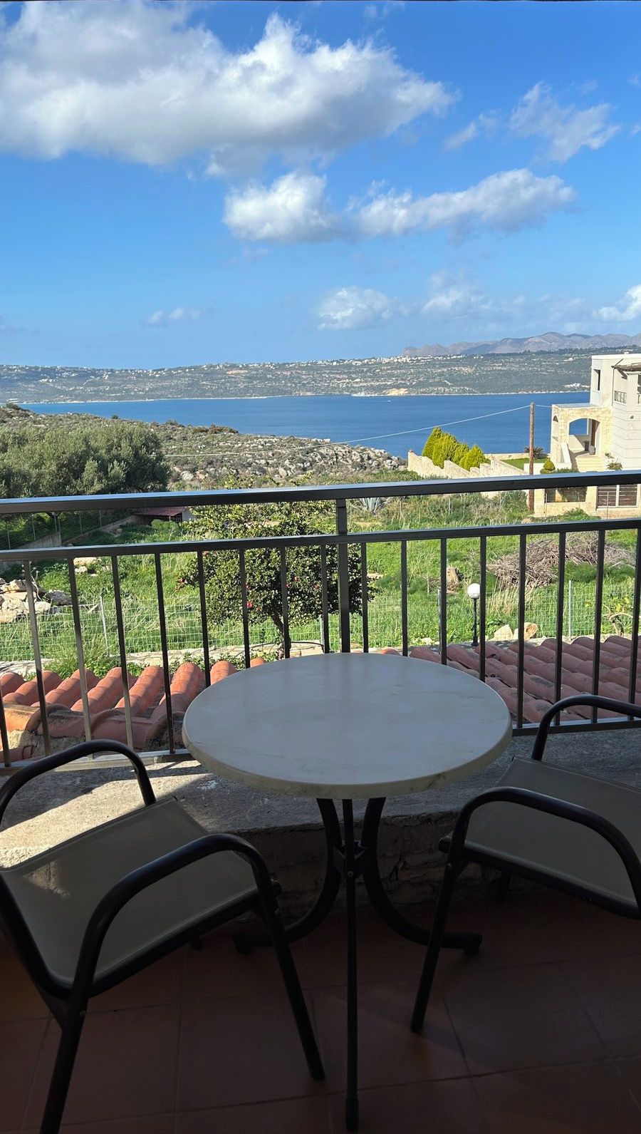 A round table with four chairs on a balcony overlooking a coastal landscape with mountains in the distance.