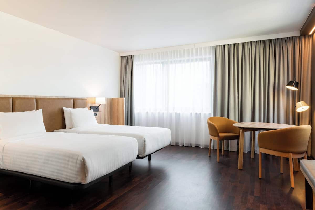 A hotel room with two beds, a wooden headboard, a small table with chairs, and a large window with curtains.