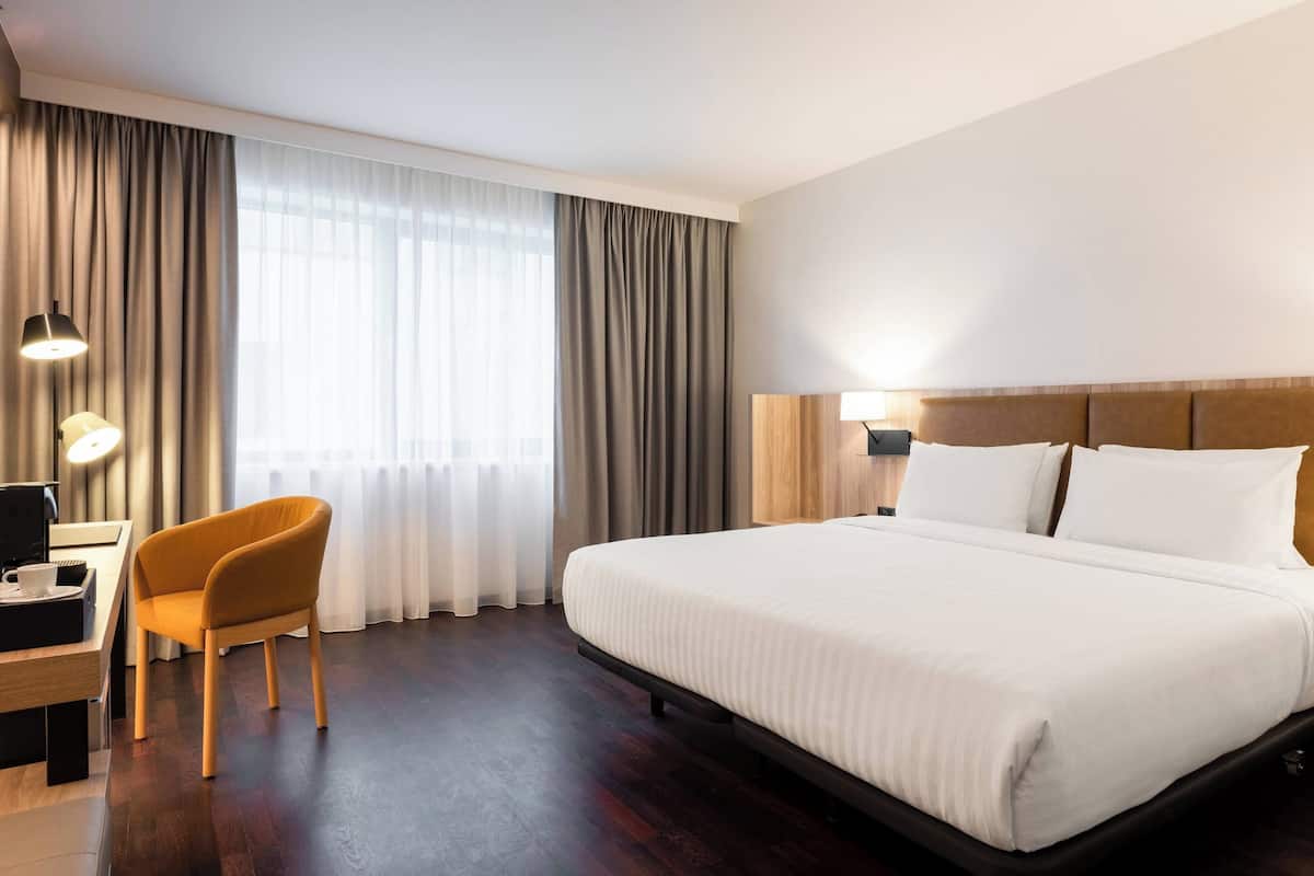 A hotel room with a large bed, a desk with a chair, a lamp, and a window with curtains.