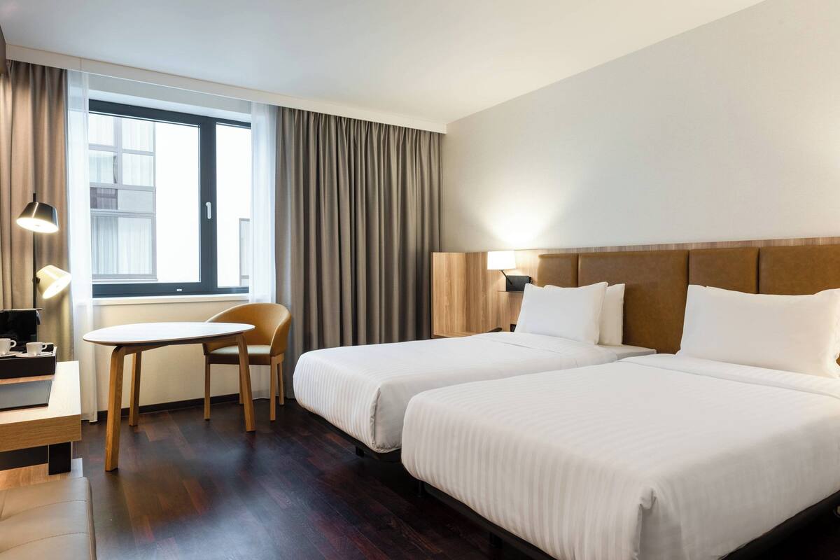 A hotel room with two beds, a wooden table, a chair, and a window with curtains.