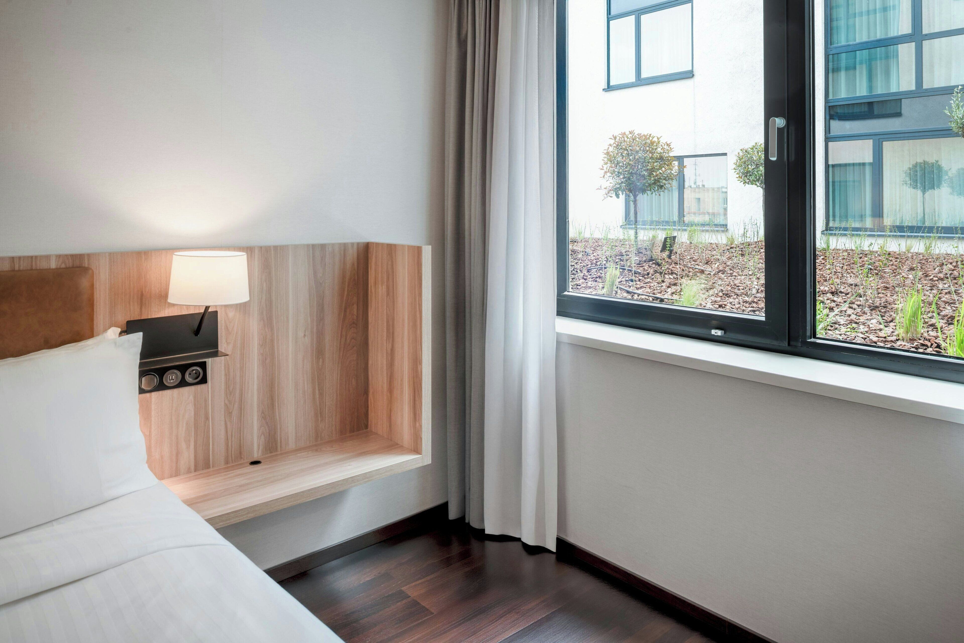 Habitación de hotel con cama, mesita de noche con lámpara, ventana con cortinas y vistas al exterior.