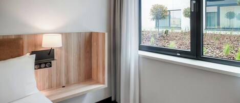 Habitación de hotel con cama, mesita de noche con lámpara, ventana con cortinas y vistas al exterior.
