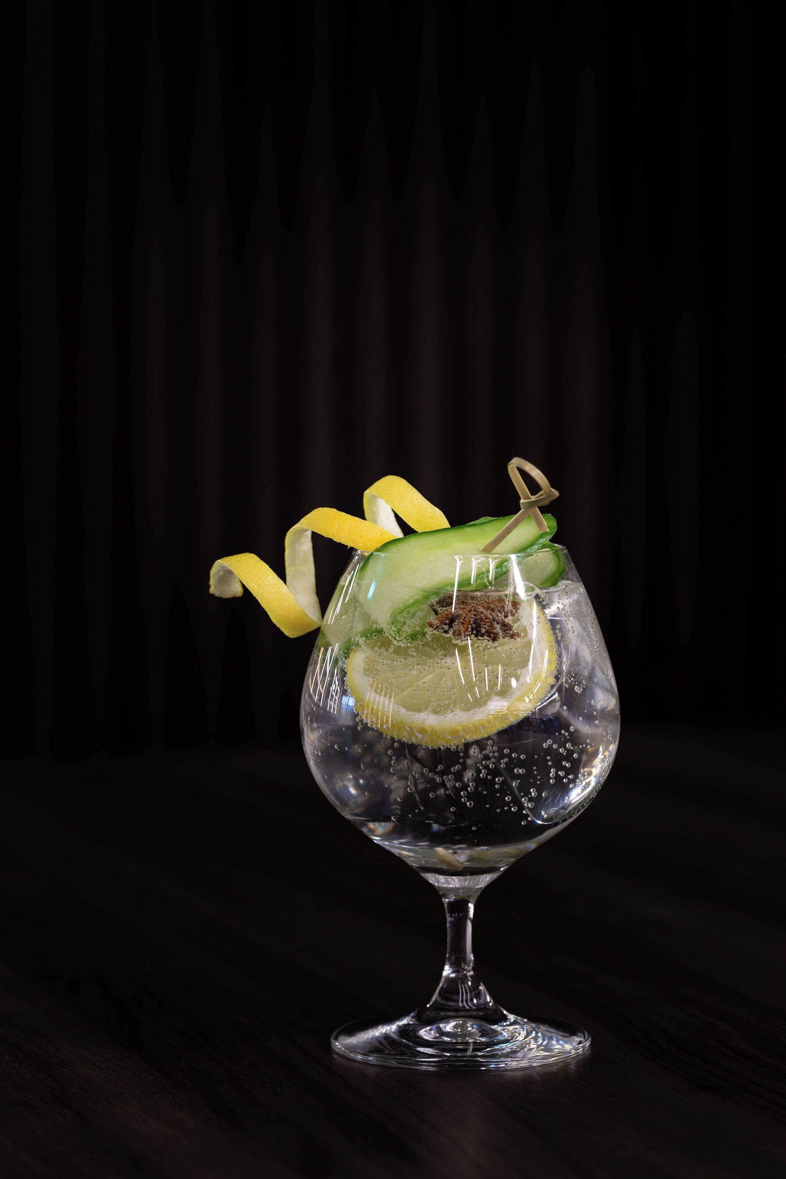A clear glass with a lime slice, cucumber, and lemon peel garnish on a dark wooden surface.