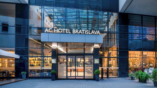 The exterior of AC Hotel Bratislava with a modern glass facade, outdoor seating area, and visible interior lighting. - AC Hotel by Marriott Bratislava Old Town (Bratislava)