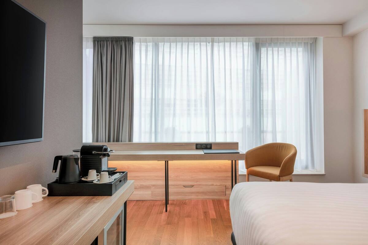 A modern hotel room with a wooden desk, a comfortable armchair, a bed, and a large window with curtains.