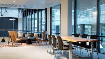 Una sala de reuniones moderna con una larga mesa, sillas, un sofá y grandes ventanales que ofrecen vistas del paisaje urbano.