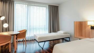 Quarto de hotel com piso de madeira, uma cama, uma mesa com duas cadeiras, uma luminária e uma janela grande com cortinas.
