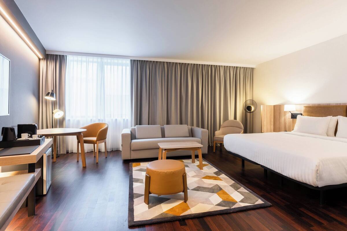 A hotel room with a bed, a sofa, a desk with a chair, a small table, and a round ottoman.