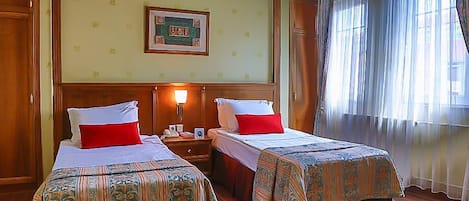 A hotel room with two beds, wooden headboards, a nightstand with a lamp, a framed picture on the wall, and a window with curtains.