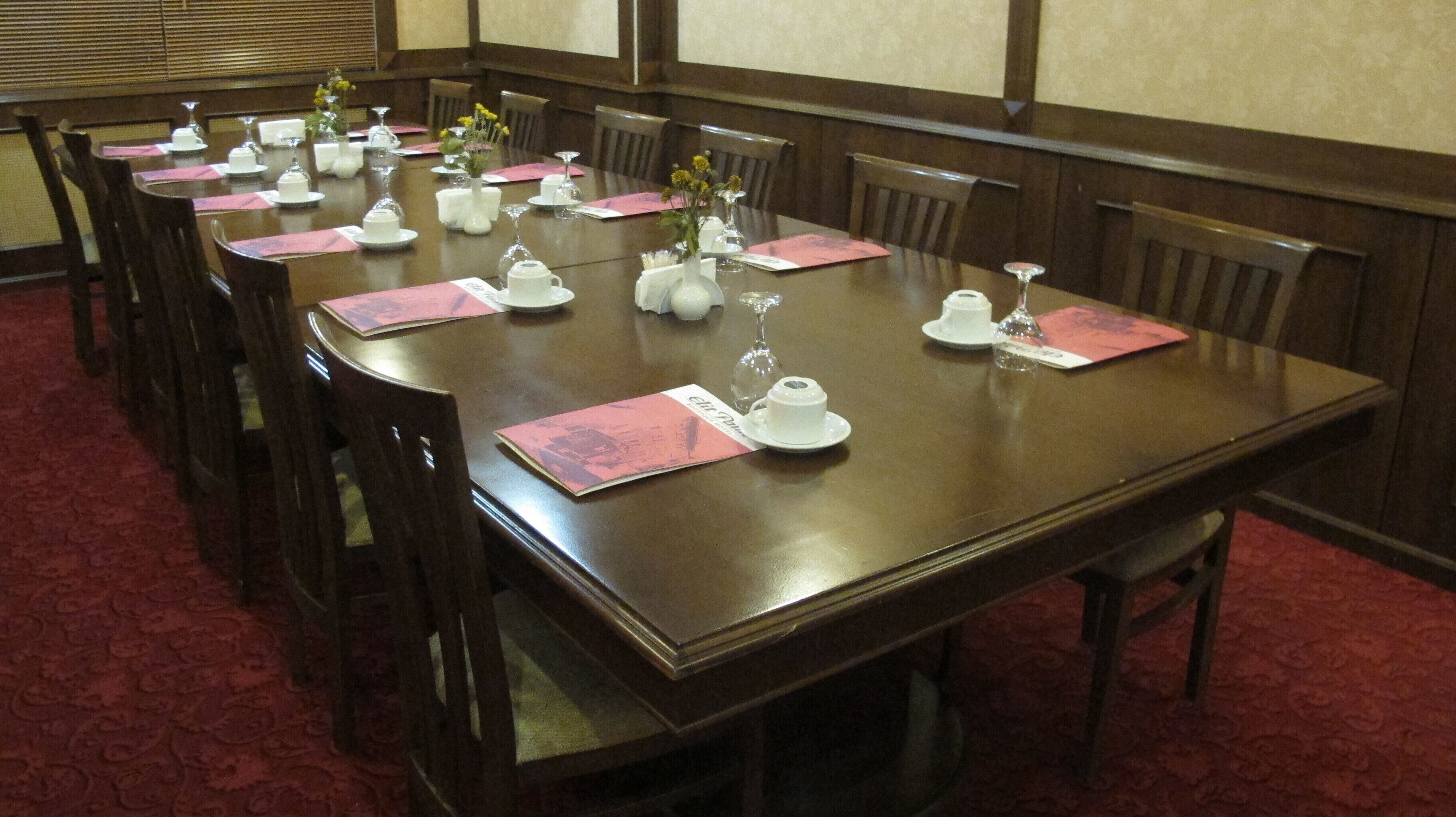 Un salón de conferencias con una larga mesa dispuesta para una reunión, equipado con tarjetas de identificación, vasos de agua y un jarrón con flores.
