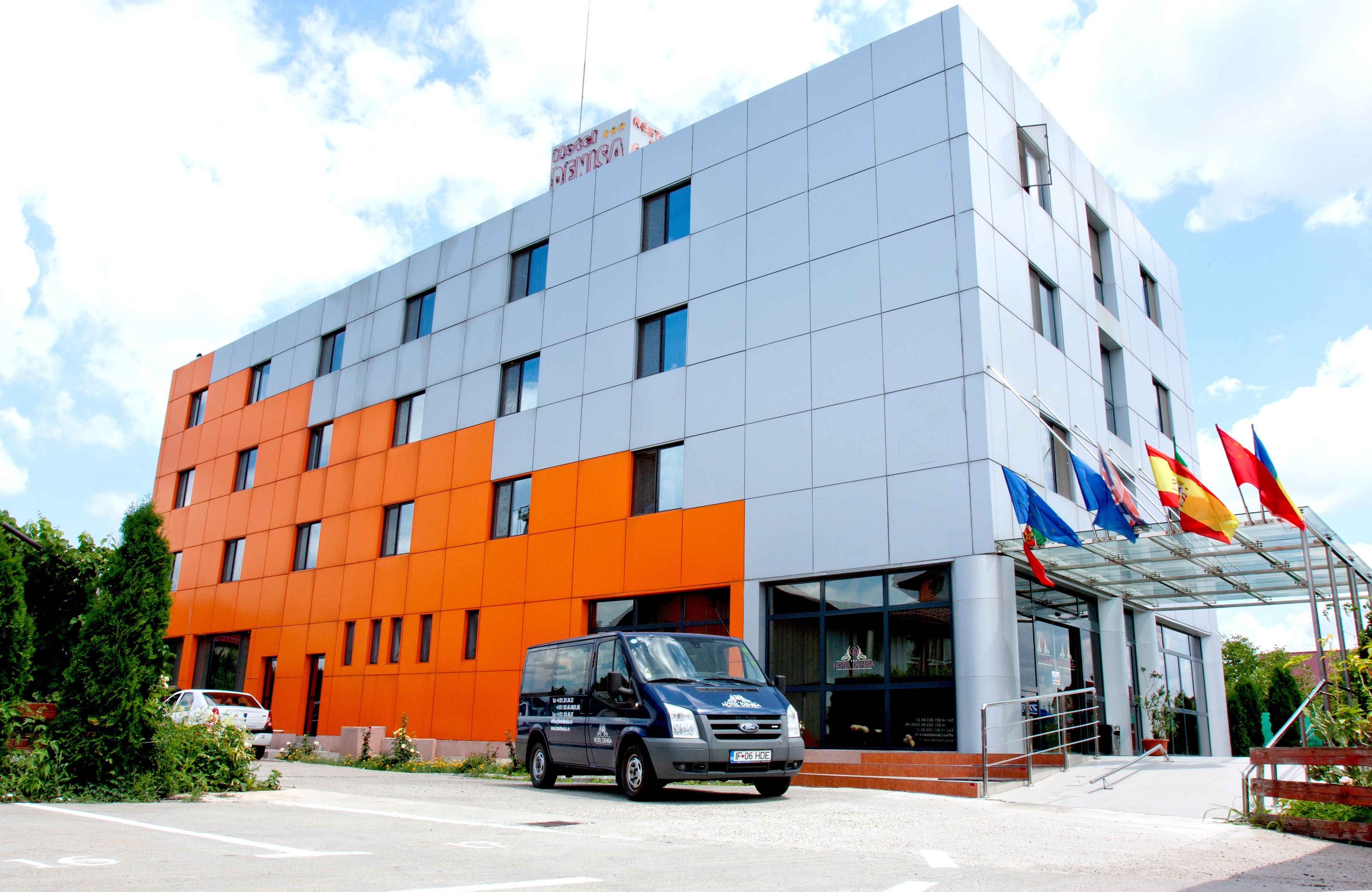 A modern building with a mix of orange and grey panels, a glass entrance, and multiple flags.