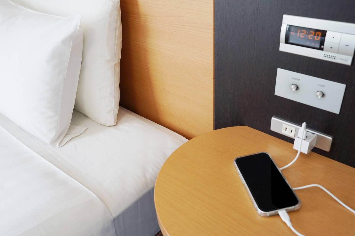 Ein Hotelzimmer mit Bett, Nachttisch, lädendem Handy, digitaler Uhr und Steckdose.