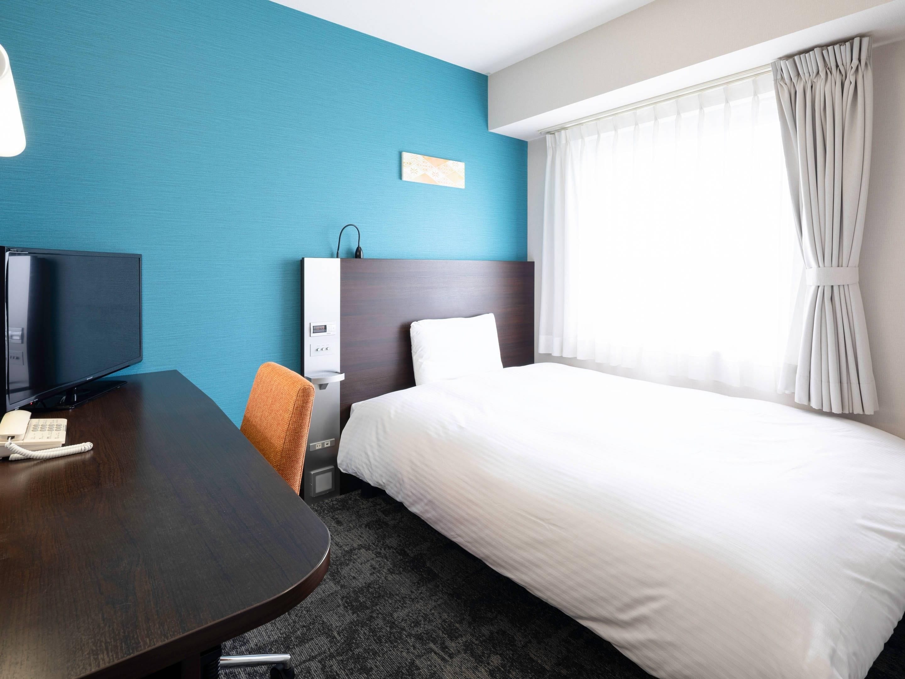 A hotel room with a bed, a desk with a telephone, a TV, and a wall-mounted panel.