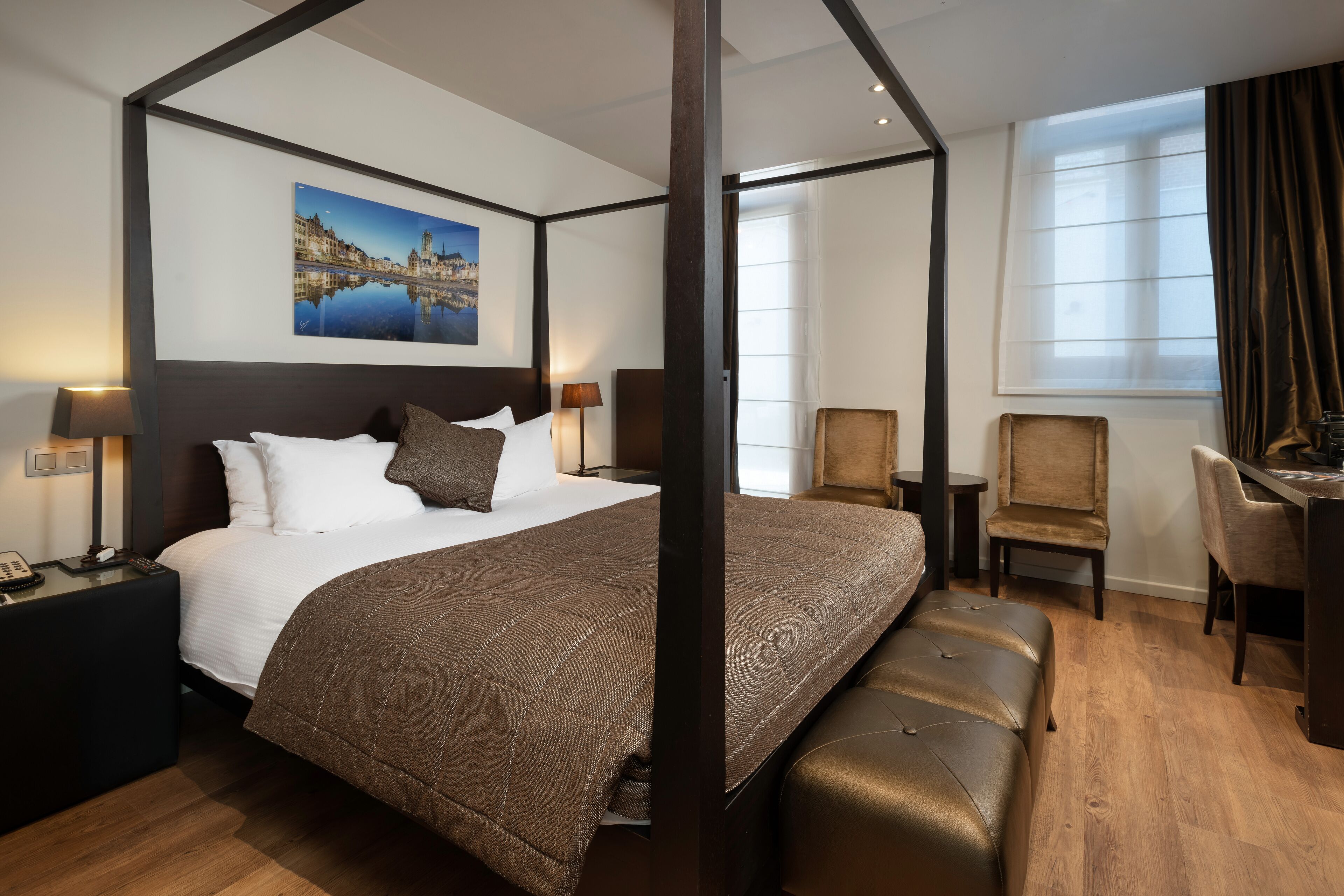 A four-poster bed with a cityscape painting, a desk with two chairs, and a large window with curtains.