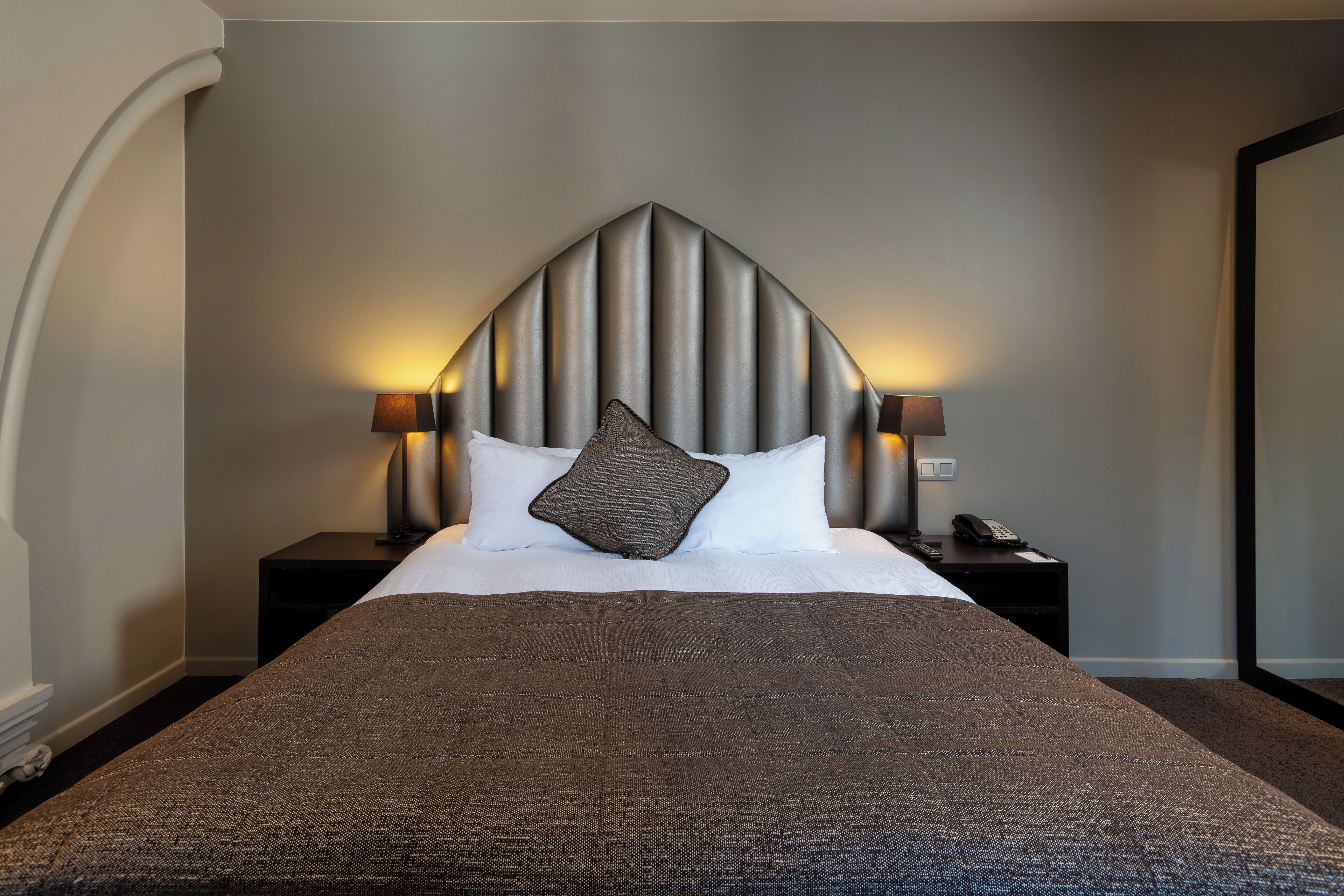 A hotel room with a large bed, a headboard with vertical lines, two bedside lamps, a telephone on the nightstand, and a dark-colored throw blanket.