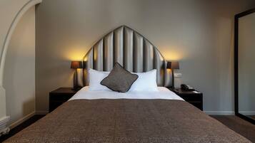 A hotel room with a large bed, a headboard with vertical lines, two bedside lamps, a telephone on the nightstand, and a dark-colored throw blanket.