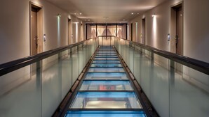 Un pasillo moderno de hotel con un ascensor de vidrio, varias puertas y una barandilla central de vidrio.