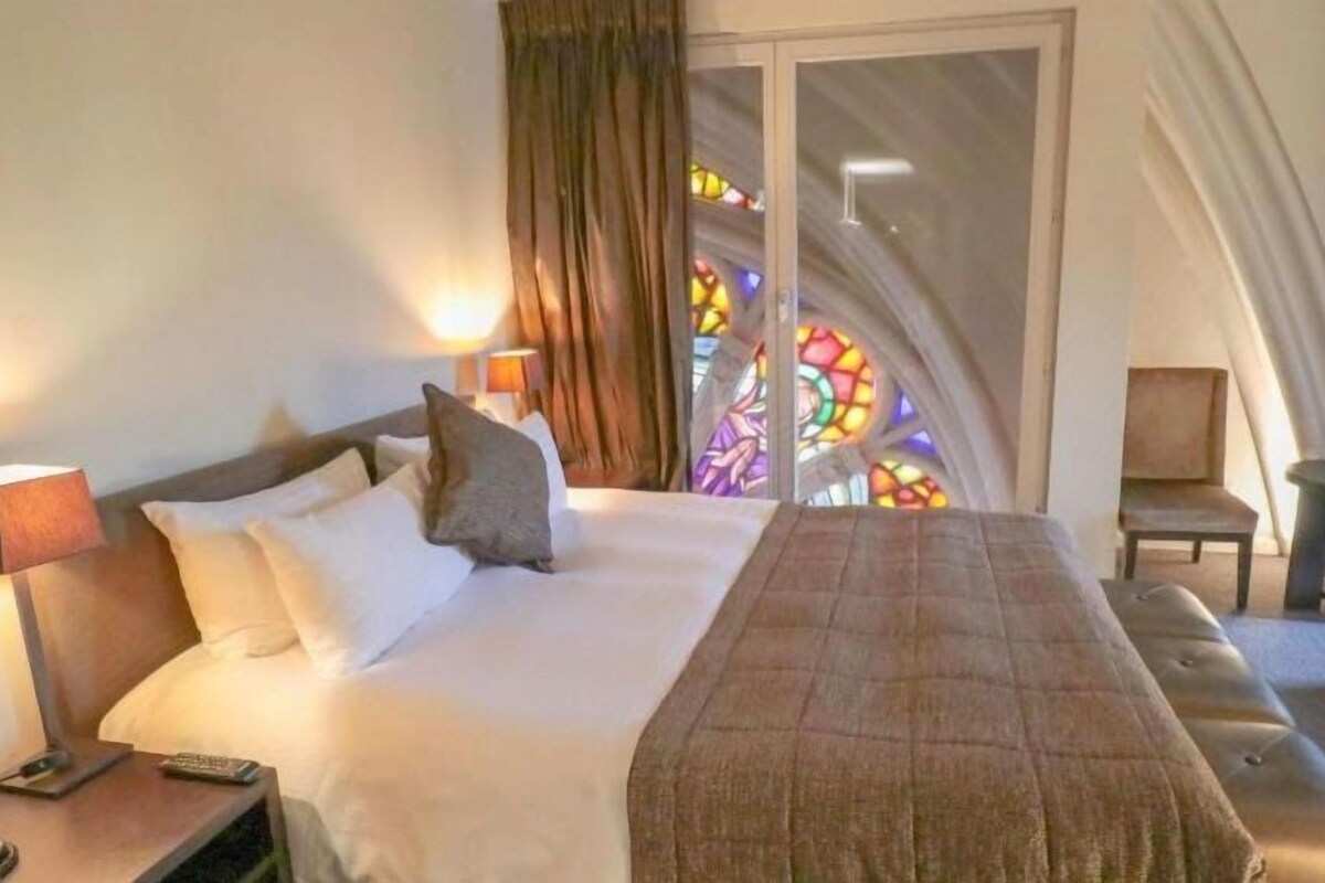 Una habitación de hotel con cama, mesita de noche, una silla y un ventanal de vitrales.