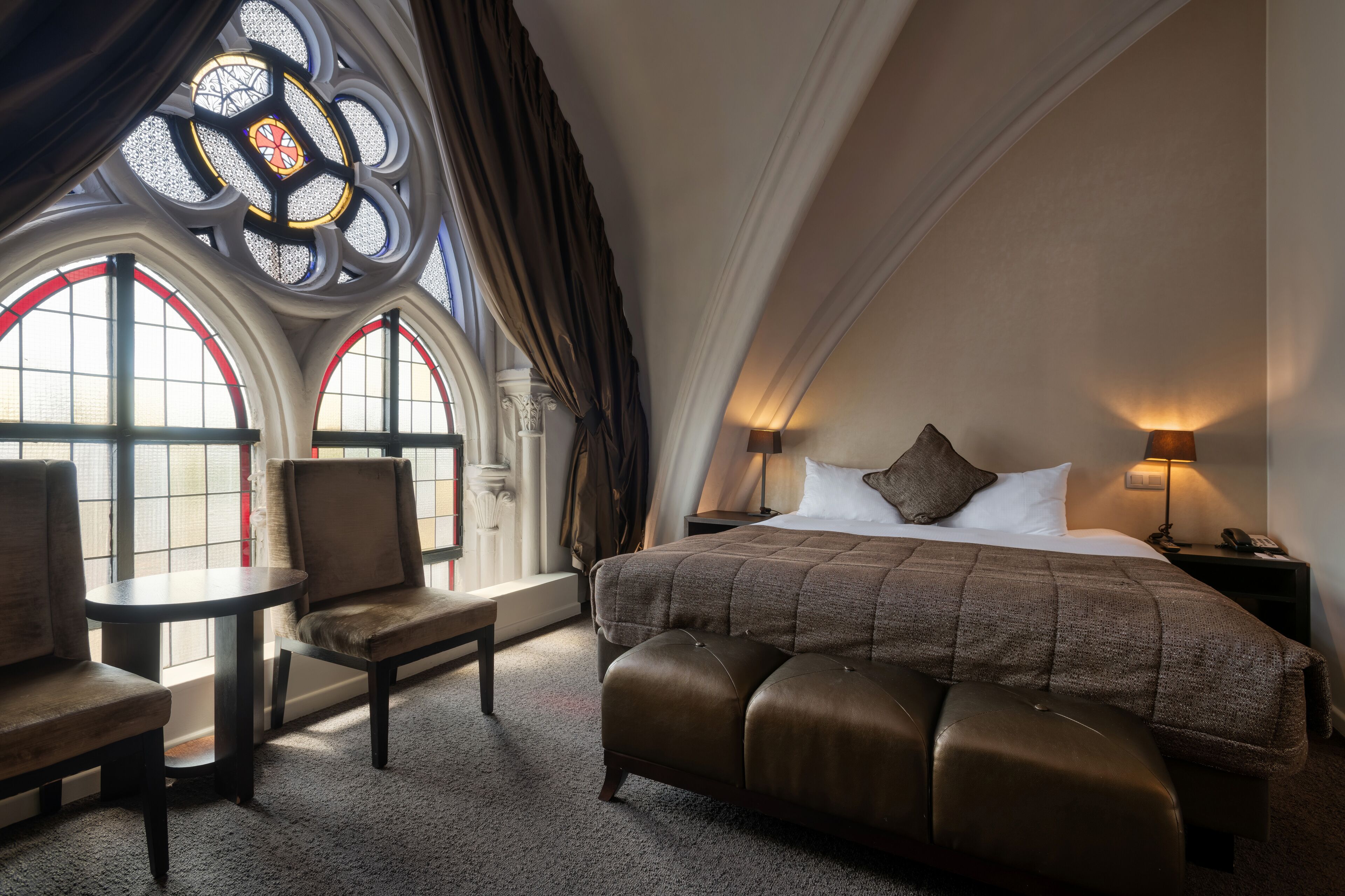 A bedroom with a large bed, a chair, a small table, and a window with stained glass.