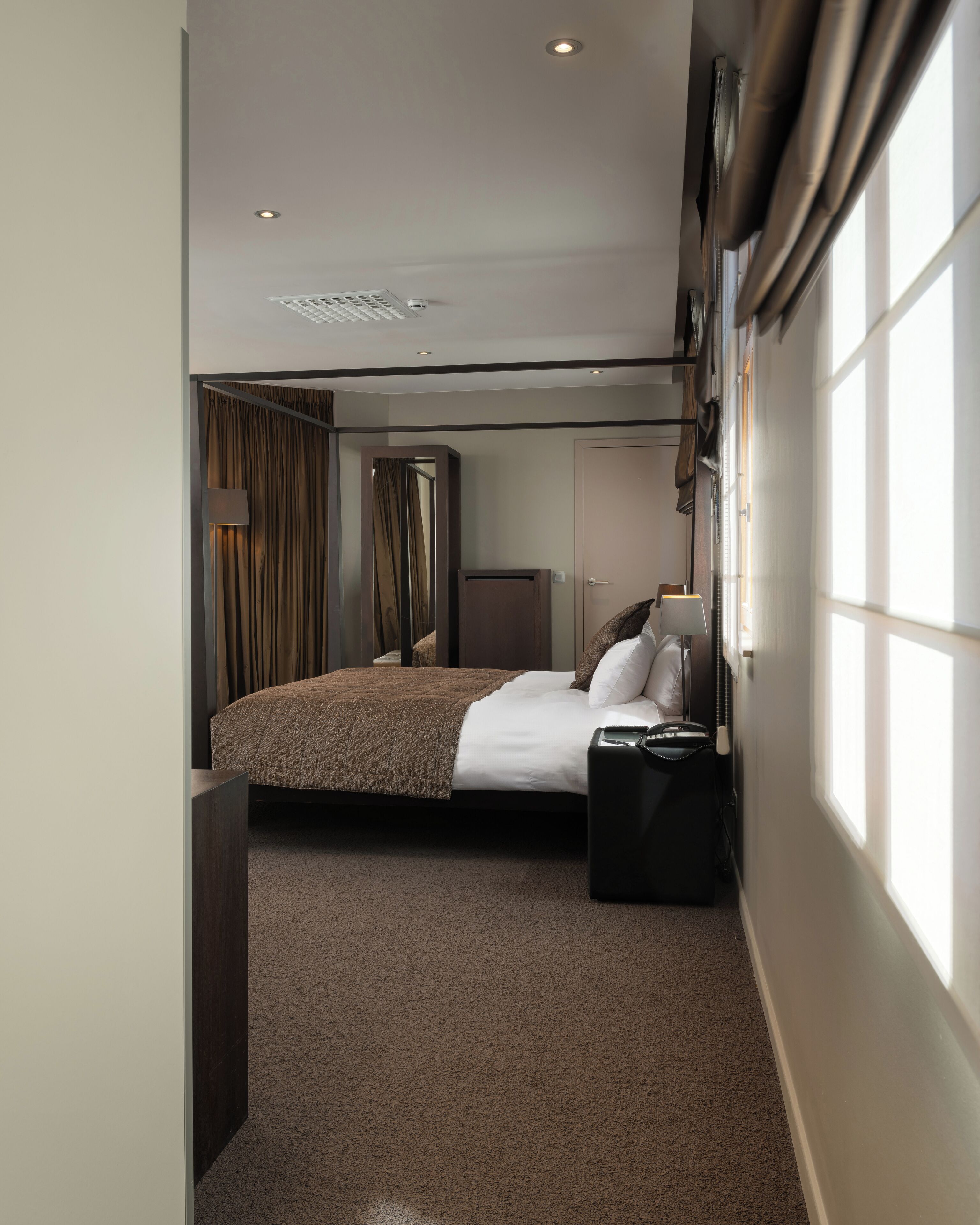 Habitación de hotel con cama, mesita de noche, lámpara, armario y ventana con cortinas.