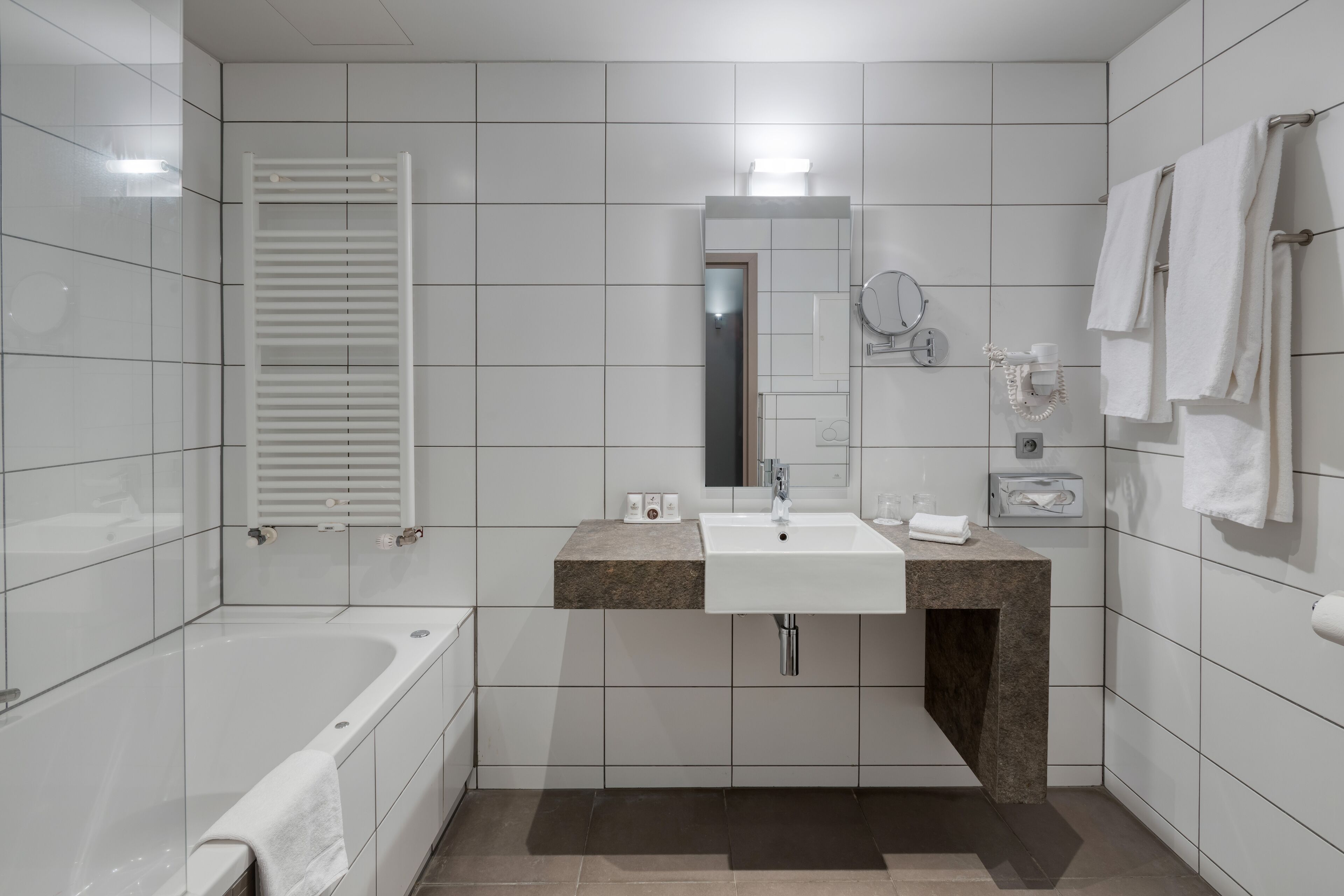 Un baño moderno con baldosas blancas, una bañera, un lavamanos con encimera de granito, un espejo y un perchero para toallas.