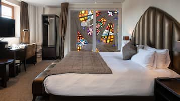 A hotel room with a large bed, a desk with a computer, a TV, and a view of a stained glass window.