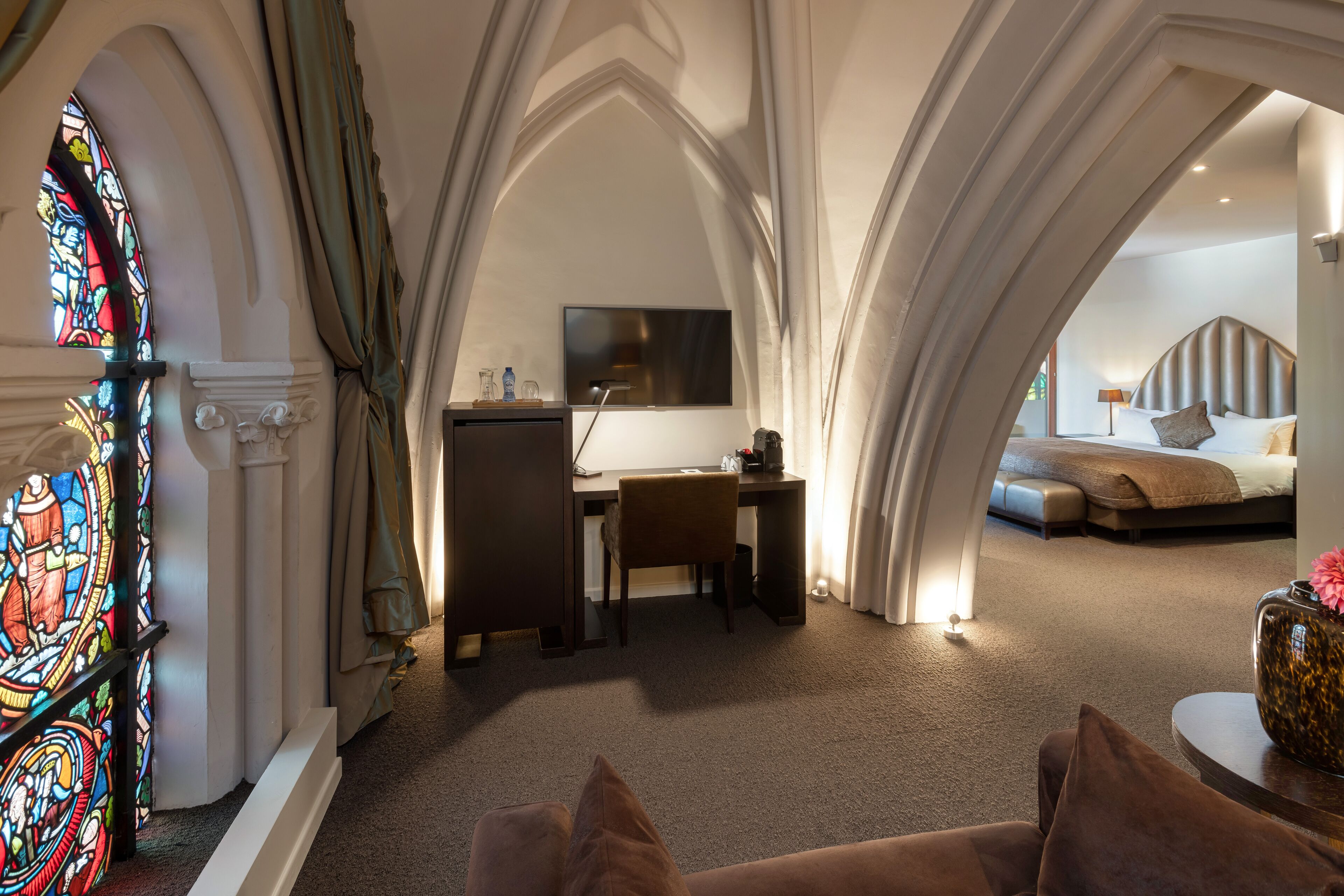 Een kamer met een bureau, stoel en een bed met hoofdeinde. Er is een glas-in-loodraam en een gordijn.
