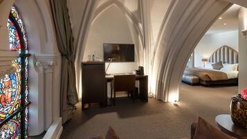 Een kamer met een bureau, stoel en een bed met hoofdeinde. Er is een glas-in-loodraam en een gordijn.