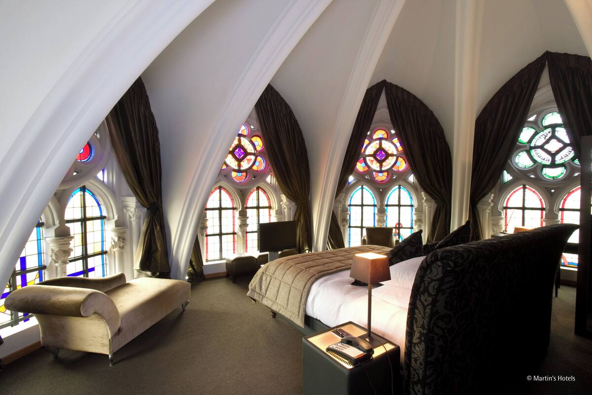 Un dormitorio con una cama grande, una silla y un escritorio con una lámpara. Se observan vidrieras y cortinas.