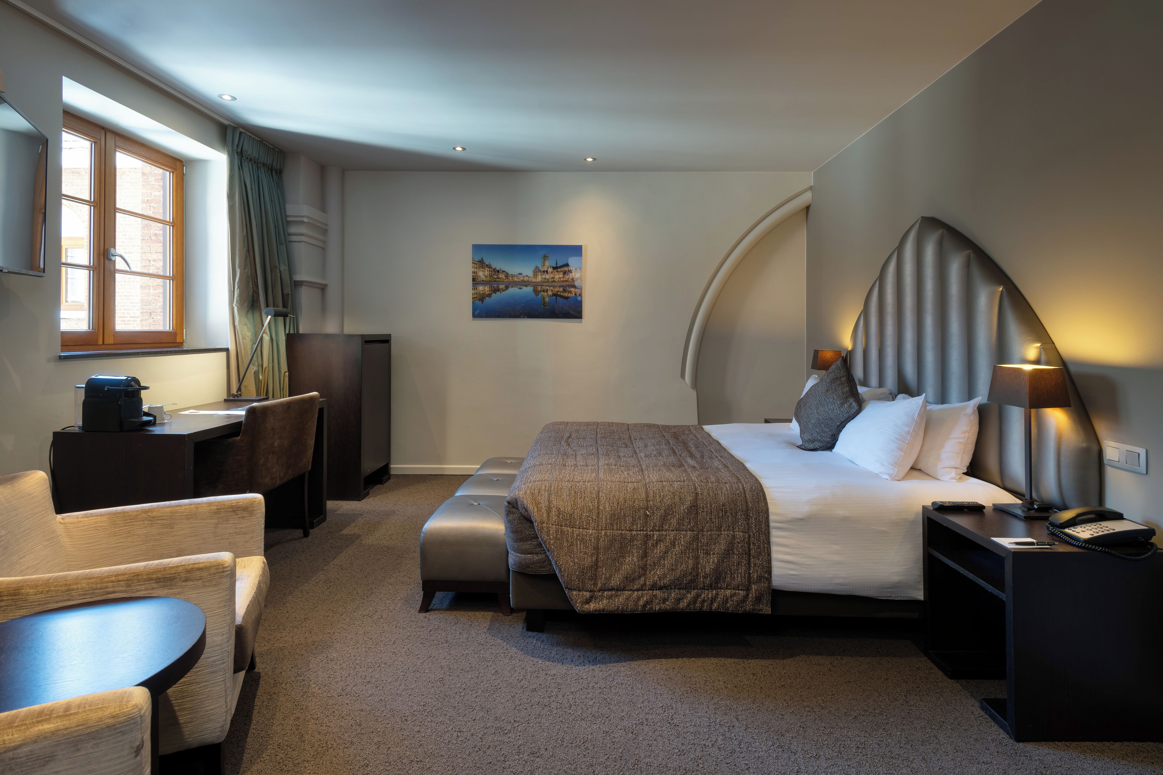 Moderni hotellihuone, jossa on sänky, yöpöytä, lamppu ja työpöytä tuolilla.