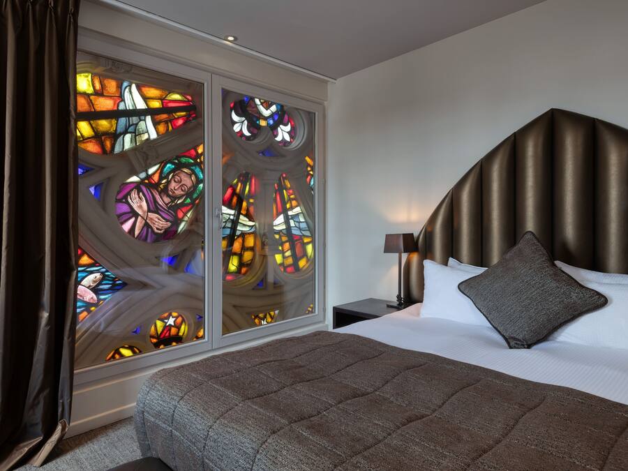 Quarto com uma cama grande, cabeceira, mesa de cabeceira e janela de vitrais.