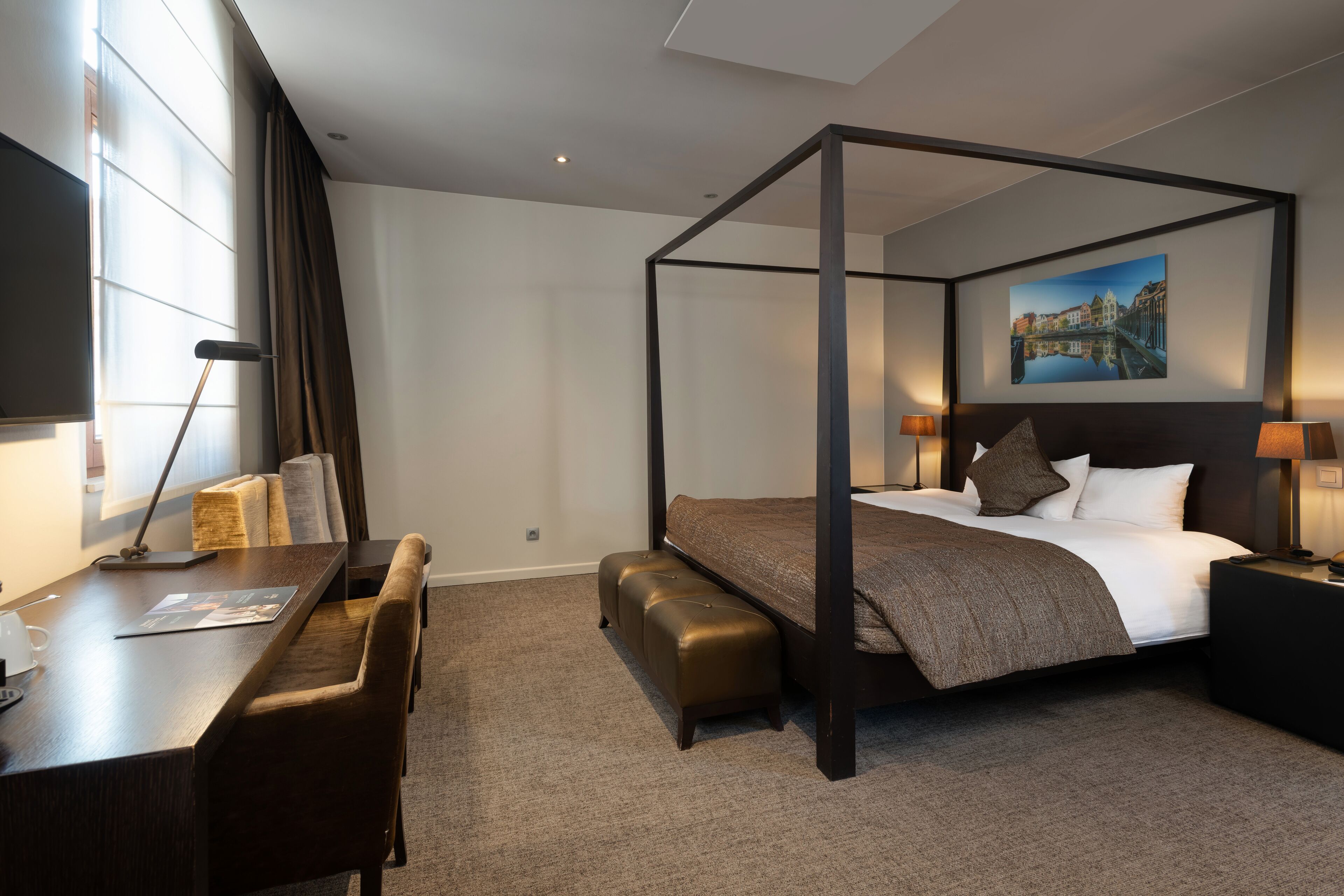 A four-poster bed with a headboard, a desk with a chair, a bench, and a nightstand with a lamp.