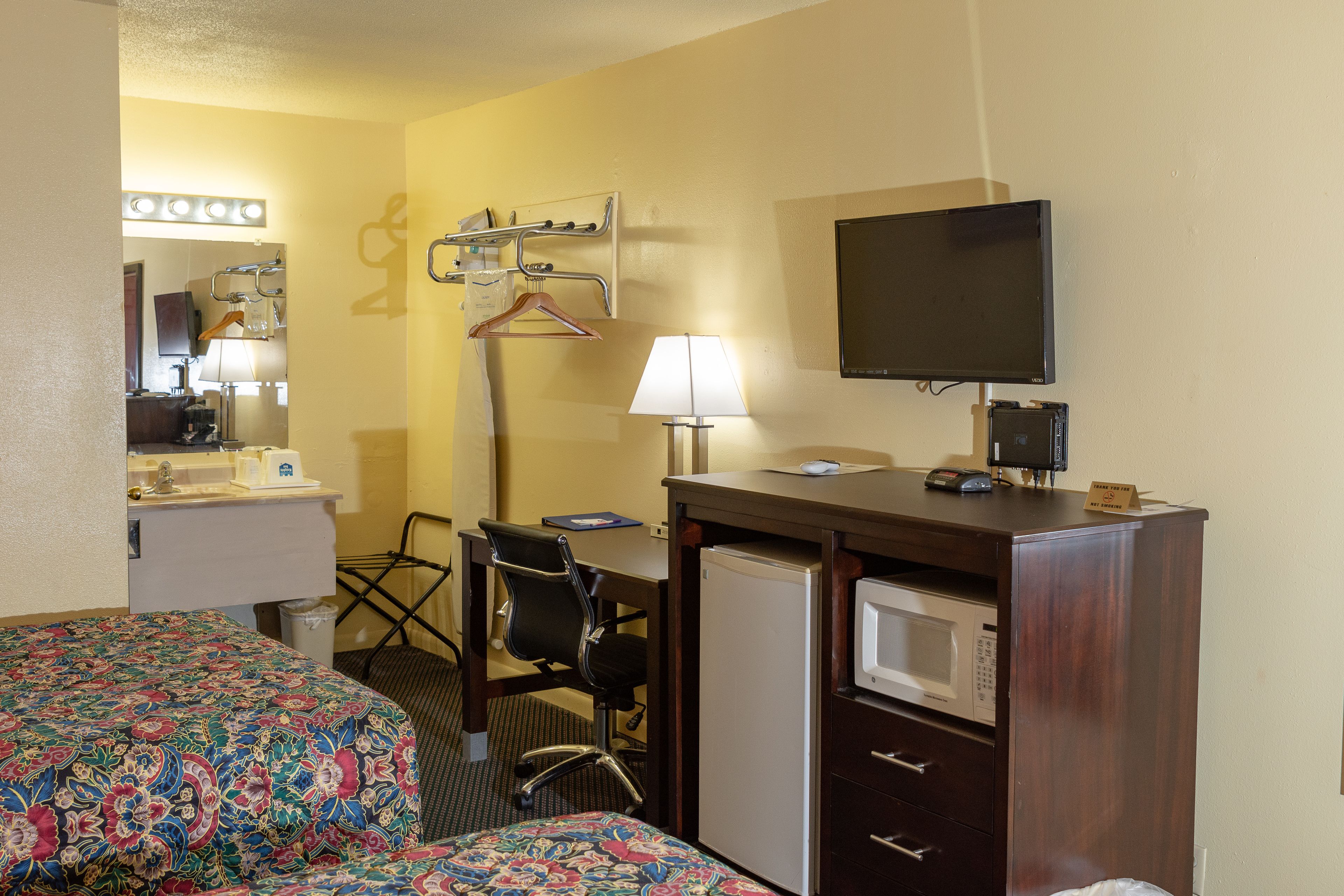 A hotel room with two beds, a desk, a chair, a desk lamp, a television, a microwave, and a mirror with lights.