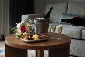 A round wooden table with a bottle of champagne, a glass of champagne, a plate of cheese and crackers, and a bouquet of flowers.