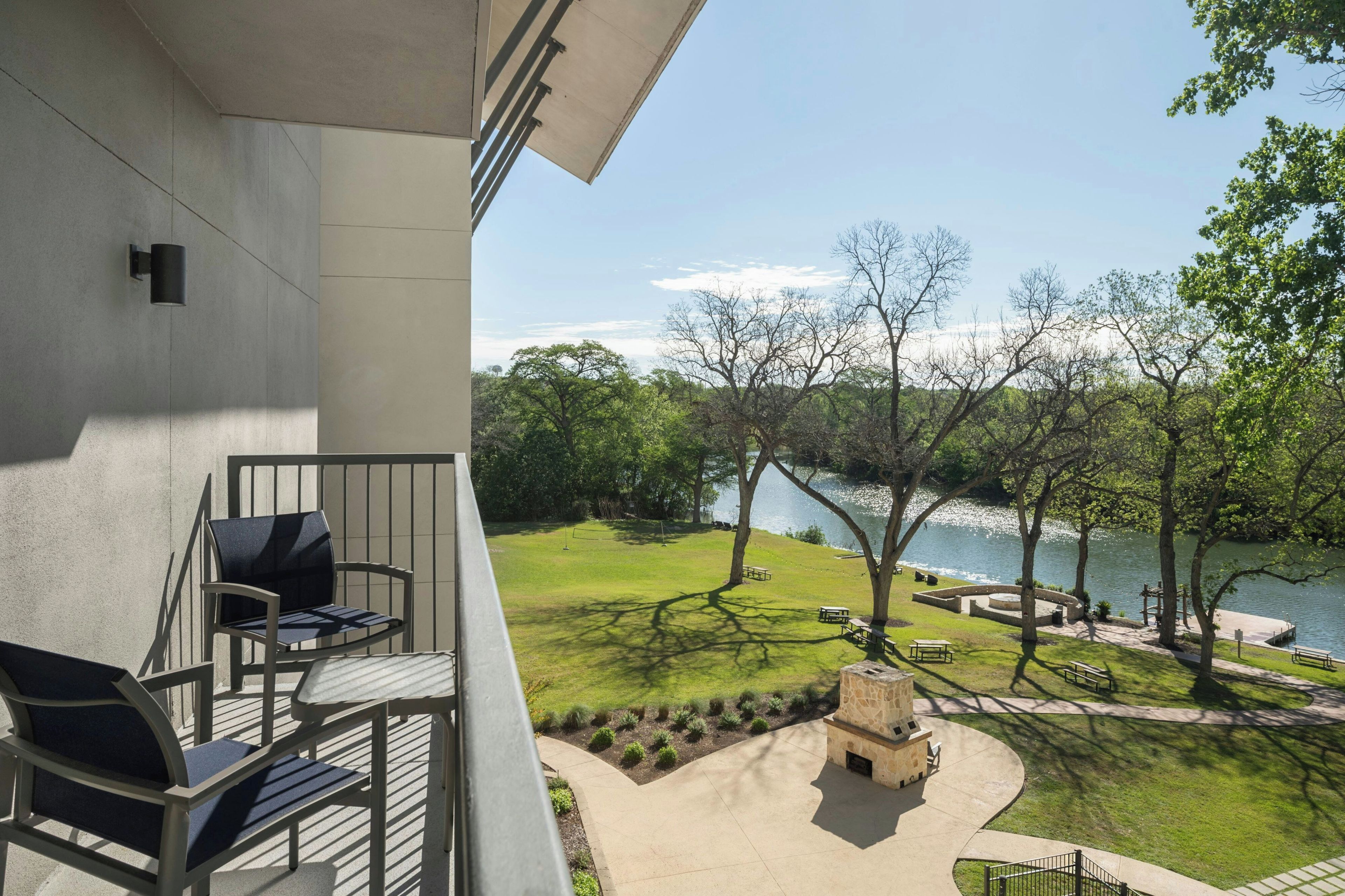A balcony with outdoor furniture, a stone fire pit, and a view of a lake surrounded by trees.