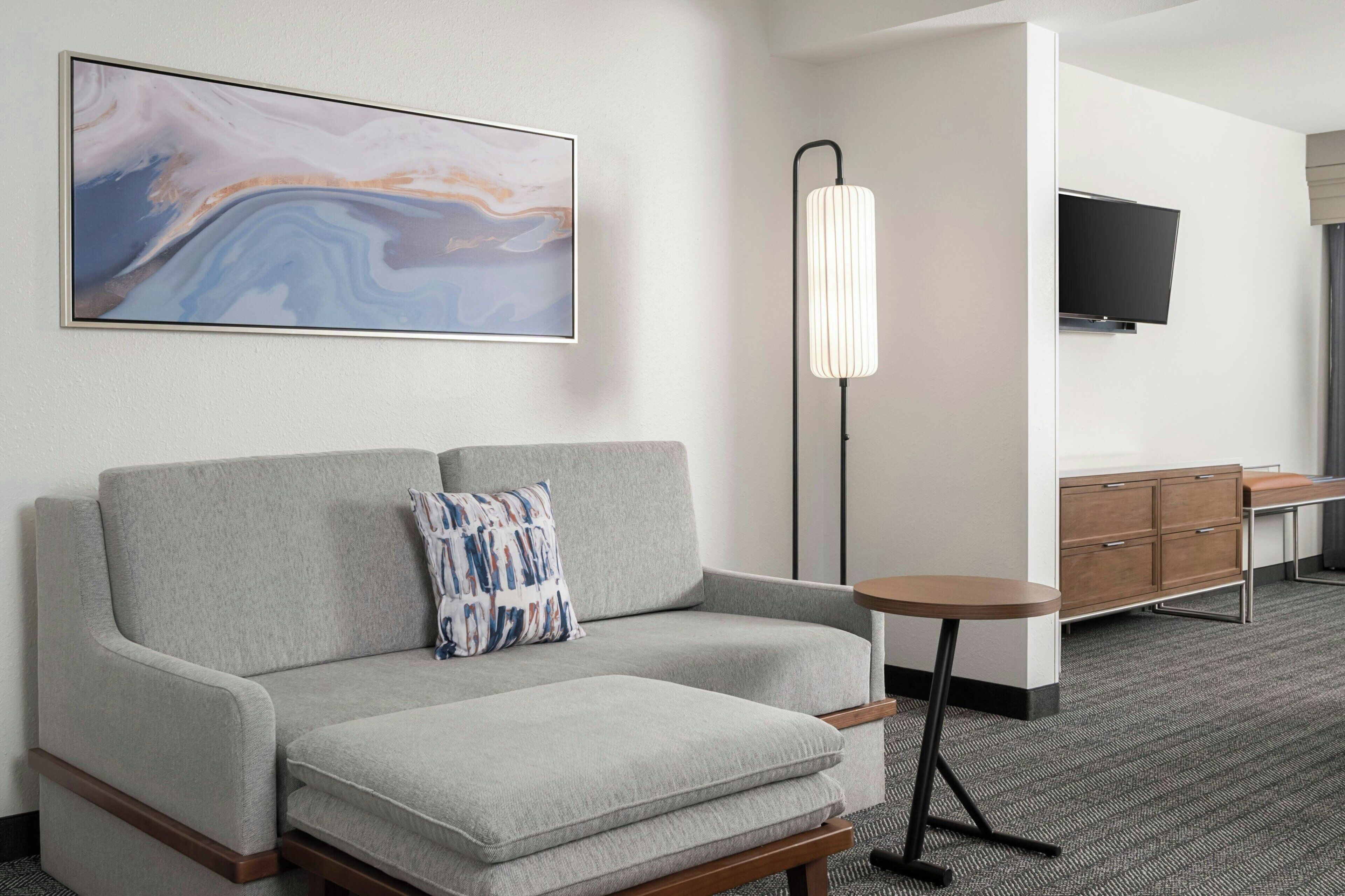 Quarto de hotel moderno com sofá cinzento, uma mesinha redonda, luminária de chão, televisão fixada na parede e um armário de madeira.