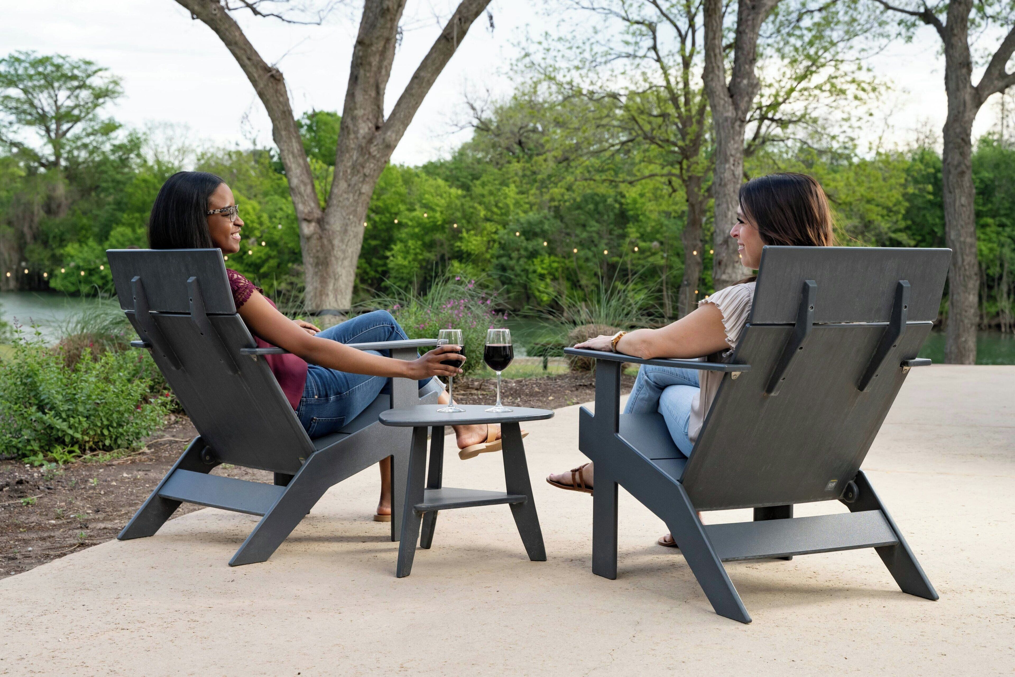 Dos personas sentadas en una mesa redonda pequeña con bebidas, al aire libre con árboles y una masa de agua al fondo.