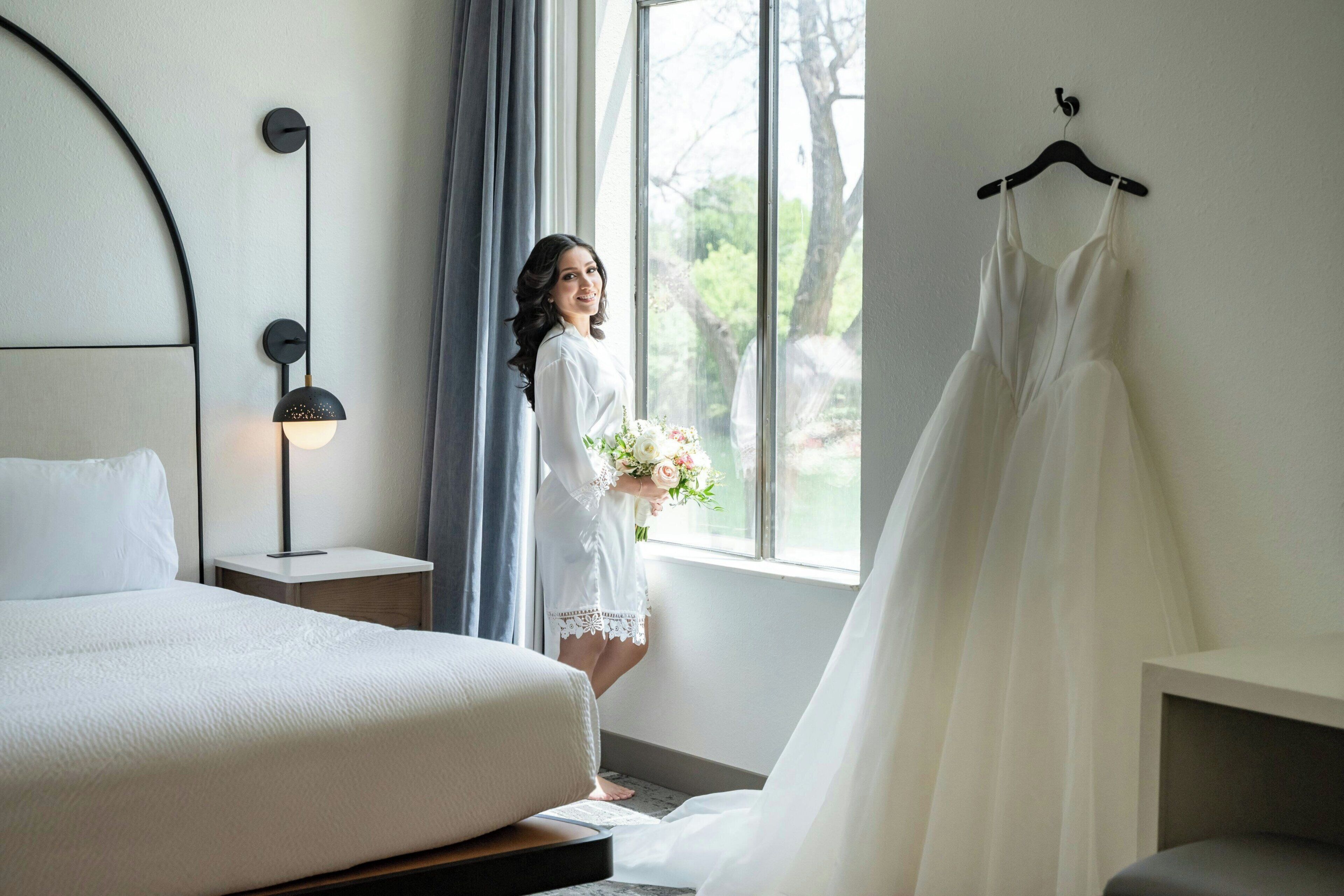 Ein Schlafzimmer mit einem Bett, einem Nachttisch, einer Wandlampe, einem Fenster mit Vorhängen und einem Kleid an einem Kleiderbügel.