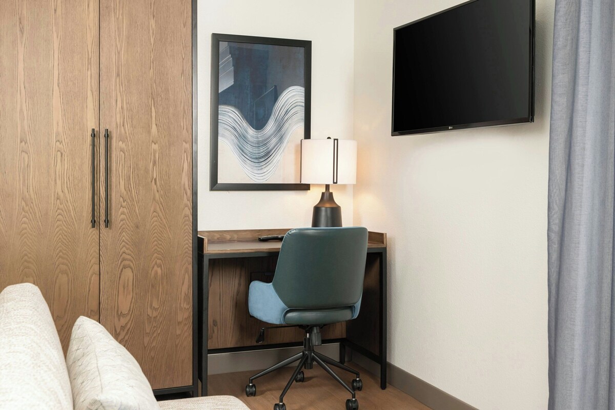 A hotel room with a wooden wardrobe, a desk with a chair, a flat-screen TV, and a lamp.