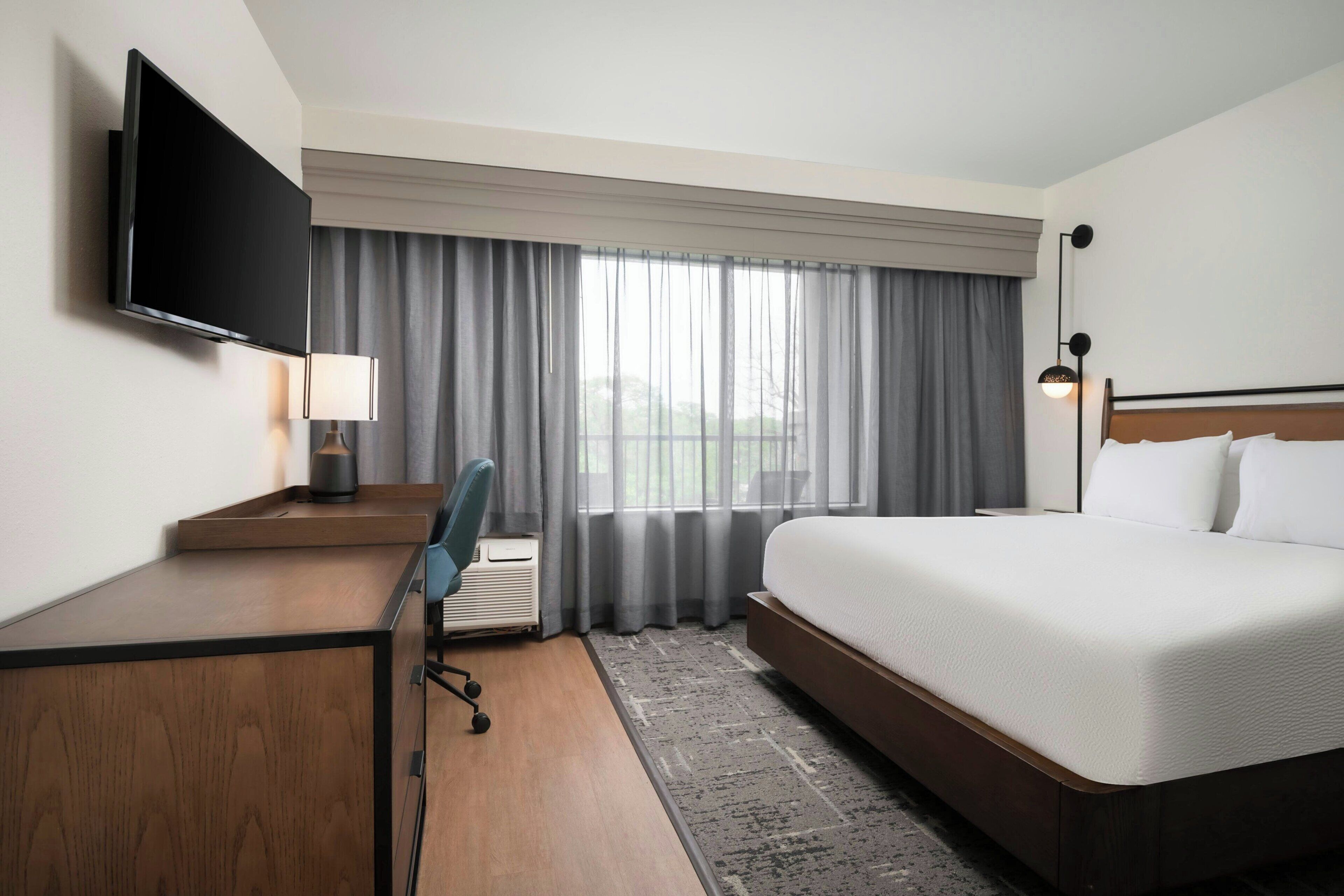 Una habitación de hotel con cama, un escritorio con silla, un televisor montado en la pared y una ventana con cortinas.