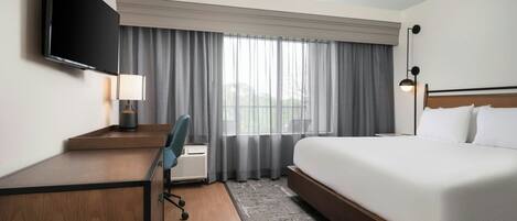 Una habitación de hotel con cama, un escritorio con silla, un televisor montado en la pared y una ventana con cortinas.