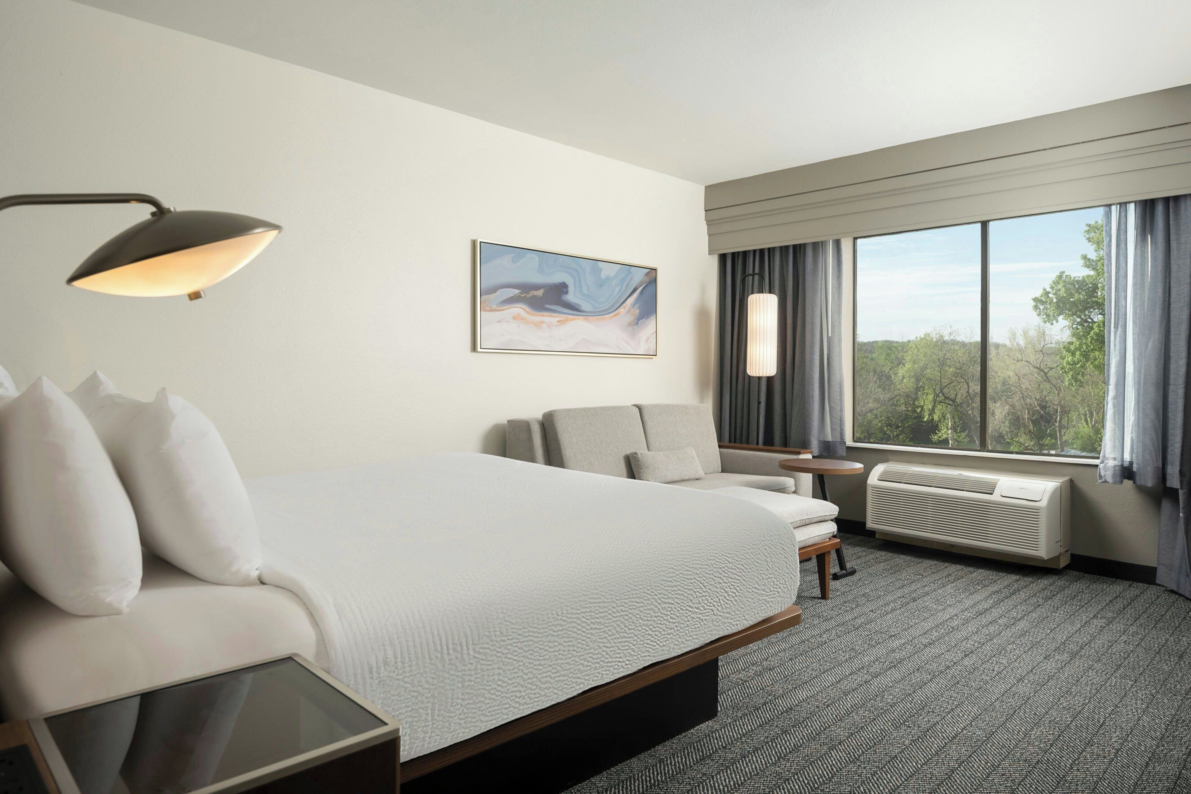 A hotel room with a bed, a desk, a chair, a window with curtains, a wall-mounted air conditioner, and a painting.