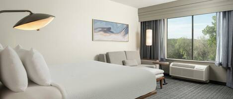 A hotel room with a bed, a desk, a chair, a window with curtains, a wall-mounted air conditioner, and a painting.