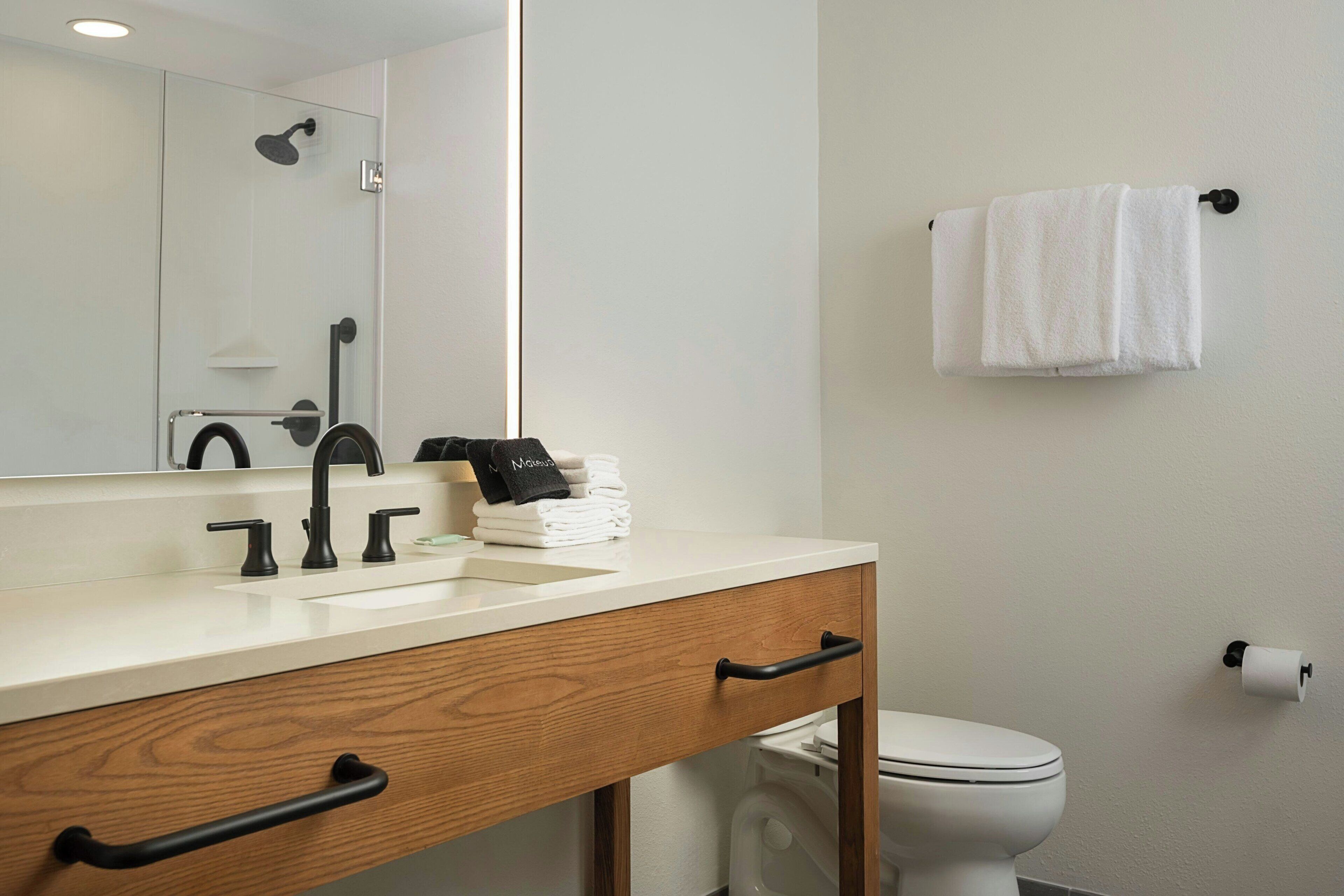 Une salle de bain avec un lavabo blanc, un robinet noir, un meuble-lavabo en bois, un miroir, un porte-serviettes et des toilettes.
