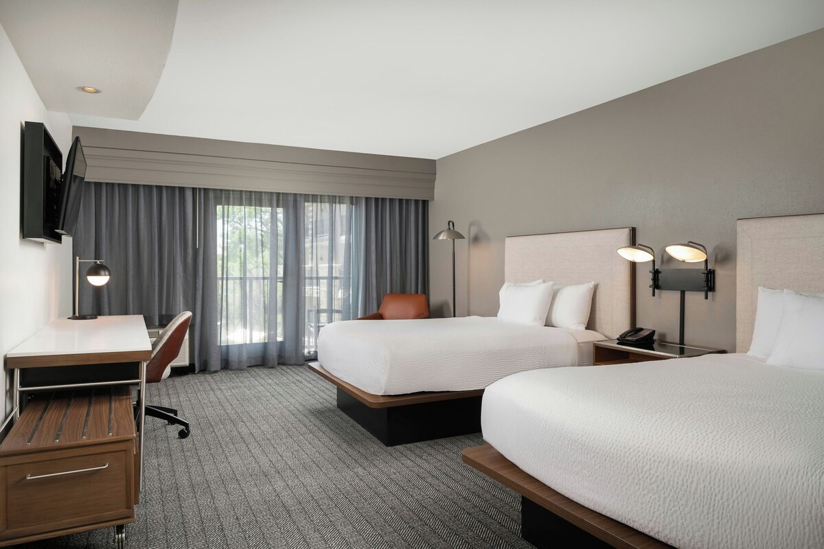 A hotel room with two beds, a desk, a chair, a TV, and a window with curtains.