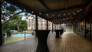 Terraza de madera al aire libre con mesas, con vistas a una piscina y un edificio residencial.